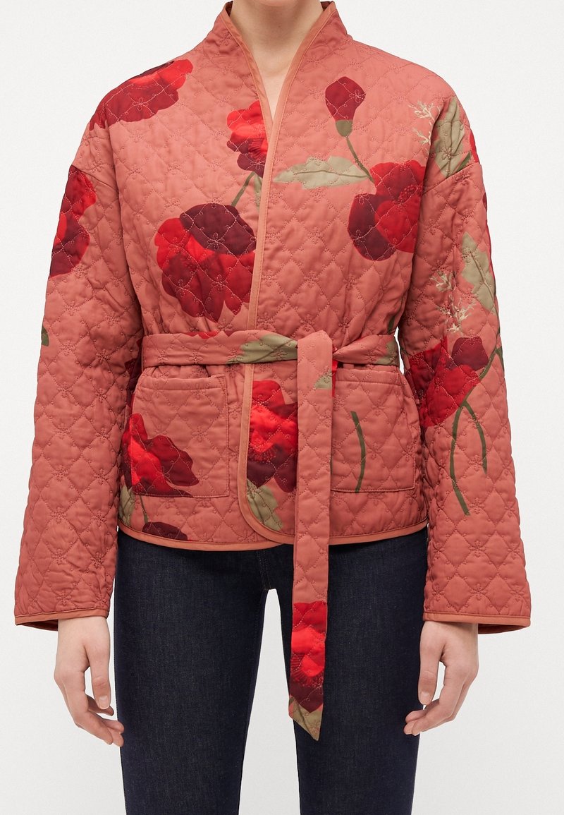 Quiltet jakke i støvet rosa med blomsterprint, bælte i taljen og sidelommer; har brede ærmer og dekorativ syning.