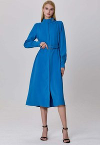 Robe longue à manches longues bleu avec un col haut, taille cintrée et une ceinture avec un accent circulaire. Texture lisse et longueur au niveau des genoux.