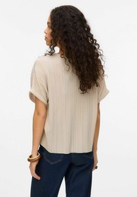 Beige plisseret bluse med korte ærmer, der viser en løs pasform. Bagpå har den en rund nederkant, og den er parret med mørke denimbukser.