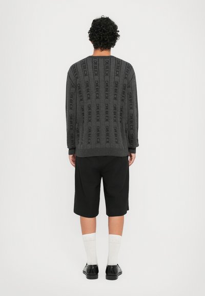 Grauer Pullover mit vertikalem Textmuster, mit gerippten Bündchen und Saum, kombiniert mit schwarzen Shorts. Weiße Socken und schwarze Schuhe runden den Look ab.