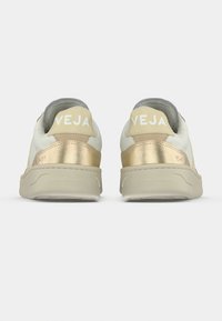 Ζευγάρι λευκά και χρυσά αθλητικά παπούτσια Veja με μπεζ σόλες, σε θέα από πίσω, εμφανίζοντας το όνομα της μάρκας στις πίσω ετικέτες.