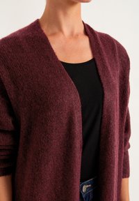 Bourgogne kofta gjord av mjukt, strukturerat tyg, med en öppen framsdesign och avkopplade ärmar, stylad över en svart topp.