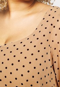 Béžová blůza vyrobená z průsvitné látky, s lodičkovým výstřihem a černými puntíky, s hladkou texturou a volným střihem.