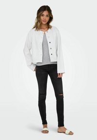 Veste en denim blanc avec un col rond et des boutons sur le devant, associée à un t-shirt gris, un jean skinny noir avec des déchirures aux genoux et des chaussures blanches à enfiler.