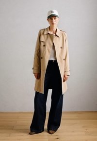 Abrigo beige con amplios solapas y botones de doble botonadura, combinado con una parte superior ajustada gris, jeans oscuros de pierna acampanada y una gorra blanca con detalles verdes.