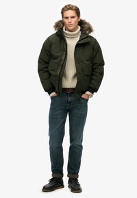 Veste matelassée verte avec capuche en fourrure, pull en maille beige, jeans bleus et bottes noires. Les accessoires comprennent une ceinture marron et une montre.