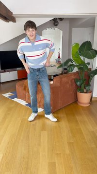 Camisa polo a rayas en azul, rojo y blanco, combinada con jeans azul claro y zapatillas blancas. De pie en un entorno moderno de sala de estar.