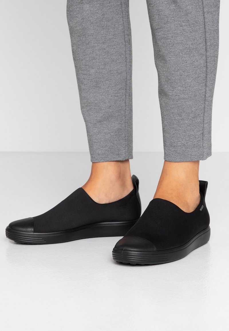 Pieds portant des baskets noires slip-on associées à un pantalon gris, debout sur un sol blanc lisse avec un fond uni.