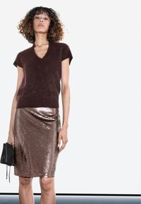 Brauner, kurzärmeliger V-Ausschnitt-Pullover kombiniert mit einem metallisch roségoldenen Pailletten-Bleistiftrock. Das Model hält eine kleine schwarze Clutch.