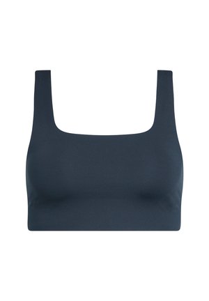 Junge Frau in schwarzem Crop-Top, schwarzen Jogginghosen und silbernen Sneakers steht mit den Händen in den Taschen und schaut nach unten.