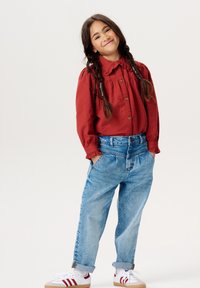 Bordowa koszula na guziki z falbaniastym kołnierzykiem, w zestawieniu z jasnoniebieskimi luźnymi dżinsami. Białe trampki z czerwonymi akcentami. Fryzura w postaci dwóch warkoczy.