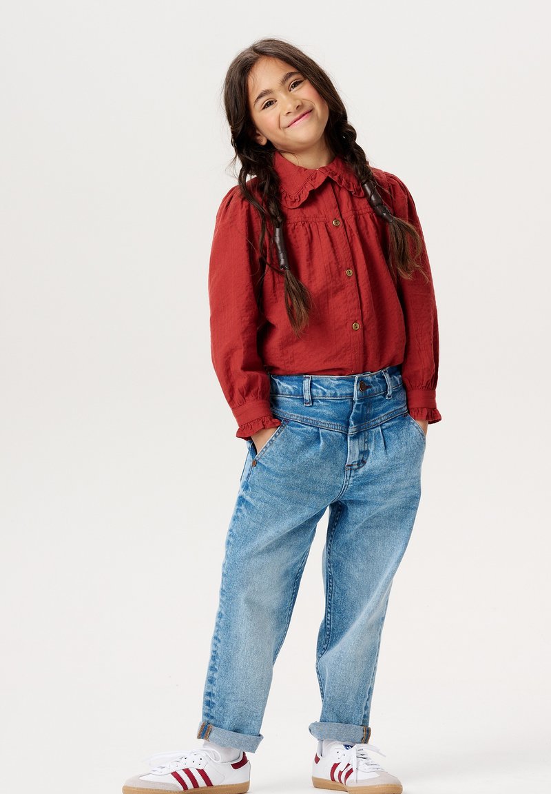 Bordowa koszula na guziki z falbaniastym kołnierzykiem, w zestawieniu z jasnoniebieskimi luźnymi dżinsami. Białe trampki z czerwonymi akcentami. Fryzura w postaci dwóch warkoczy.