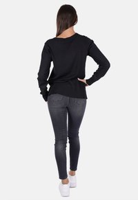 Maglione lungo nero a maniche lunghe con scollatura tonda, abbinato a jeans skinny grigi scuri e sneakers bianche. La vista posteriore mostra delle fessure laterali nel maglione.