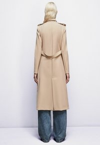 Bežový trenčkot s prispôsobeným strihom, dlhými rukávmi a zakriveným detailom pásu, kombinovaný s voľnými džínsovými nohavicami. Hladká textúra látky.