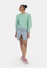 Felpa verde menta con maniche a palloncino, abbinata a una gonna di jeans chiara con spacco frontale e sneakers rosa con suola bianca.