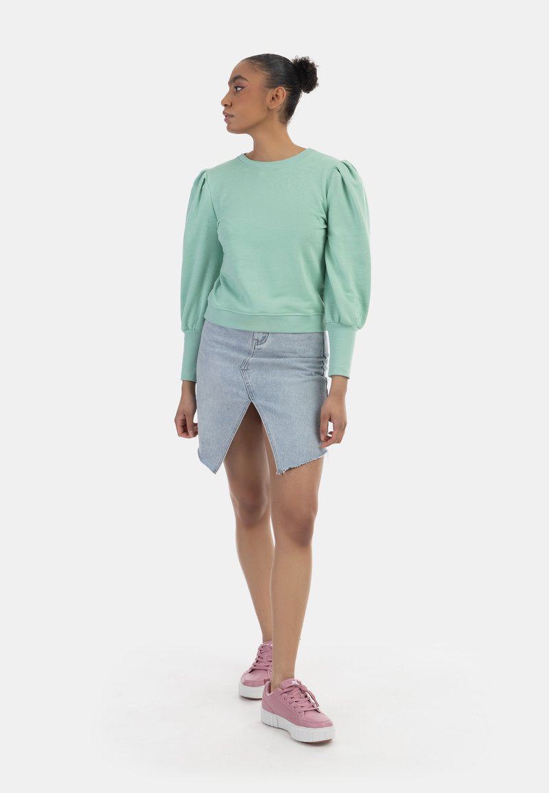 Felpa verde menta con maniche a palloncino, abbinata a una gonna di jeans chiara con spacco frontale e sneakers rosa con suola bianca.