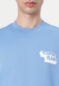 Gaiši zils kokvilnas sweatshirt. Iekļauj ribotu apkakli un baltu "TOMMY JEANS" logo kreisajā krūšu daļā. Gluda tekstūra.