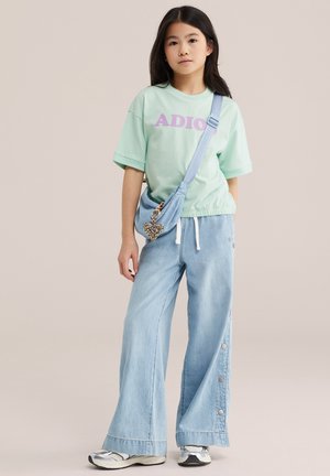 Niña joven de pie, vestida con una camisa verde menta que dice "ADIOS", pantalones anchos azul claro, zapatillas deportivas y un bolso cruzado con un charm de corazón de leopardo.