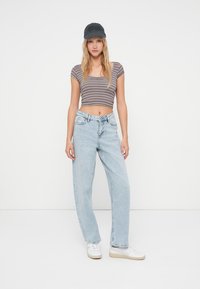 Top cropped riscado em castanho e bege, jeans de cintura alta em azul claro, tênis brancos e um boné preto com texto. Look casual, corte descontraído.