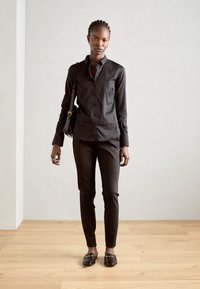 Camicia nera con bottoni abbinata a pantaloni neri slim-fit. Il modello indossa mocassini neri e porta una piccola borsa nera. Tessuto liscio, design su misura.