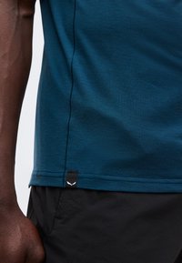 Κοντινό ενός ατόμου που φοράει ένα σκούρο τιρκουάζ πουκάμισο με μικρή μαύρη ετικέτα κοντά στο τελείωμα και μαύρο παντελόνι, εμφανίζοντας το χέρι και το πλάι του κορμού.