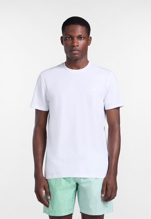 Mężczyzna stojący przodem, ubrany w prosty biały t-shirt z krótkim rękawem oraz zielone i białe szorty w pionowe paski.