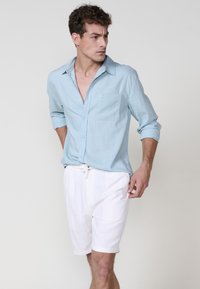 Camicia azzurro chiaro con bottoni e taschino, maniche arrotolate, abbinata a pantaloni corti bianchi con cordino. Il tessuto è morbido e ha una vestibilità rilassata.