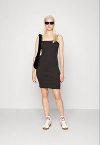 Vestido preto ajustado com decote quadrado e bordas franzidas, combinado com ténis brancos e uma mala preta de ombro felpuda. Modelo com óculos de sol.