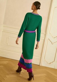 Rochie lungă verde tricotată cu guler unduit, tiv în zigzag roz și bleu, și o curea contrastantă, asortată cu cizme cu toc înalt.