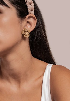 Goldene blumenförmige Ohrringe mit strukturiertem Finish, getragen am Ohr, kombiniert mit einem weißen Tanktop und einem hellen Haarband.