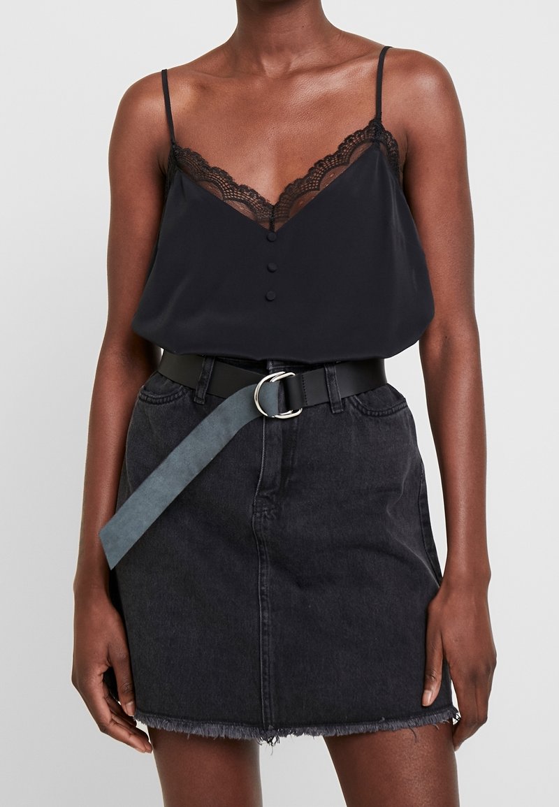 Femme portant un caraco noir orné de dentelle et une jupe en denim noire à ourlet effiloché avec une boucle de ceinture bleue lâche à l'avant.
