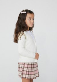 Weißer, flauschiger Cardigan mit Knopfleiste, kombiniert mit einem rosa karierten, plissierten Rock in Brauntönen, Gelb und Grün.