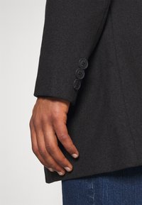 Blazer su misura in misto lana di colore grigio scuro. Presenta un colletto strutturato, quattro bottoni neri sul polsino e un orlo dritto.