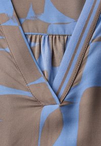 Gros plan sur un tissu floral bleu et beige avec un col en V et des plis froncés, avec une bordure texturée rayée le long du bord du col.