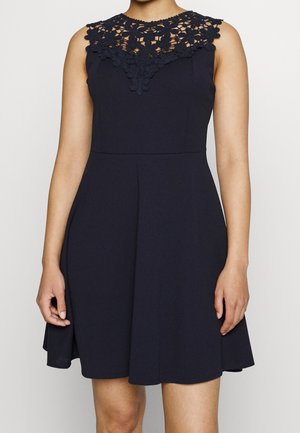 Robe noire sans manches avec détail en dentelle florale sur le décolleté, taille ajustée et jupe évasée se terminant au-dessus des genoux.