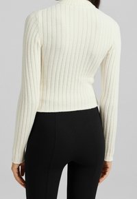 Vit ribbade tröja med hög krage, skräddarsydd design och långa ärmar, tillsammans med svarta högmidjade byxor. Mjuk textur.