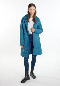 Teal quiltet frakke med hætte, der har trykknapper og lynlås. Båret over en hvid top og blå jeans, parret med sorte ankelstøvler.