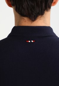 Námořnické polo tričko s texturovaným límcem; na zadním výstřihu se nachází malý vyšitý design v červené, bílé a černé barvě.