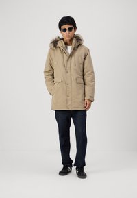 Parka beige con cappuccio bordato in pelliccia, bottoni frontali e tasche laterali. Indossata sopra jeans scuri e scarpe nere. Il modello ha i capelli corti e occhiali da sole.