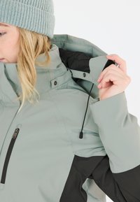 Ljusgrå utomhusjacka med svarta accenter, hög krage, justerbar dragsko och zippered ficka på bröstet. Slät textur.