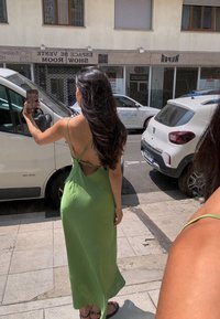 Zelena satenska maxi obleka z tankimi nastavljivimi naramnicami, globokim izrezom na hrbtu in ohlapnim krojem. Nosilka drži telefon in ujame svojo odsev.
