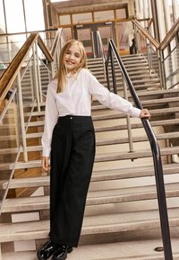 Weißes Langarmhemd kombiniert mit hoch taillierten, weiten schwarzen Hosen. Das Model trägt glänzende schwarze Schuhe und steht auf einer Innentreppe.