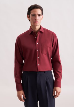Rotes Hemd mit langen Ärmeln und Knopfleiste, mit einer Brusttasche und weißen Knöpfen. Getragen mit marineblauen Hosen, der Stoff wirkt glatt und strukturiert.
