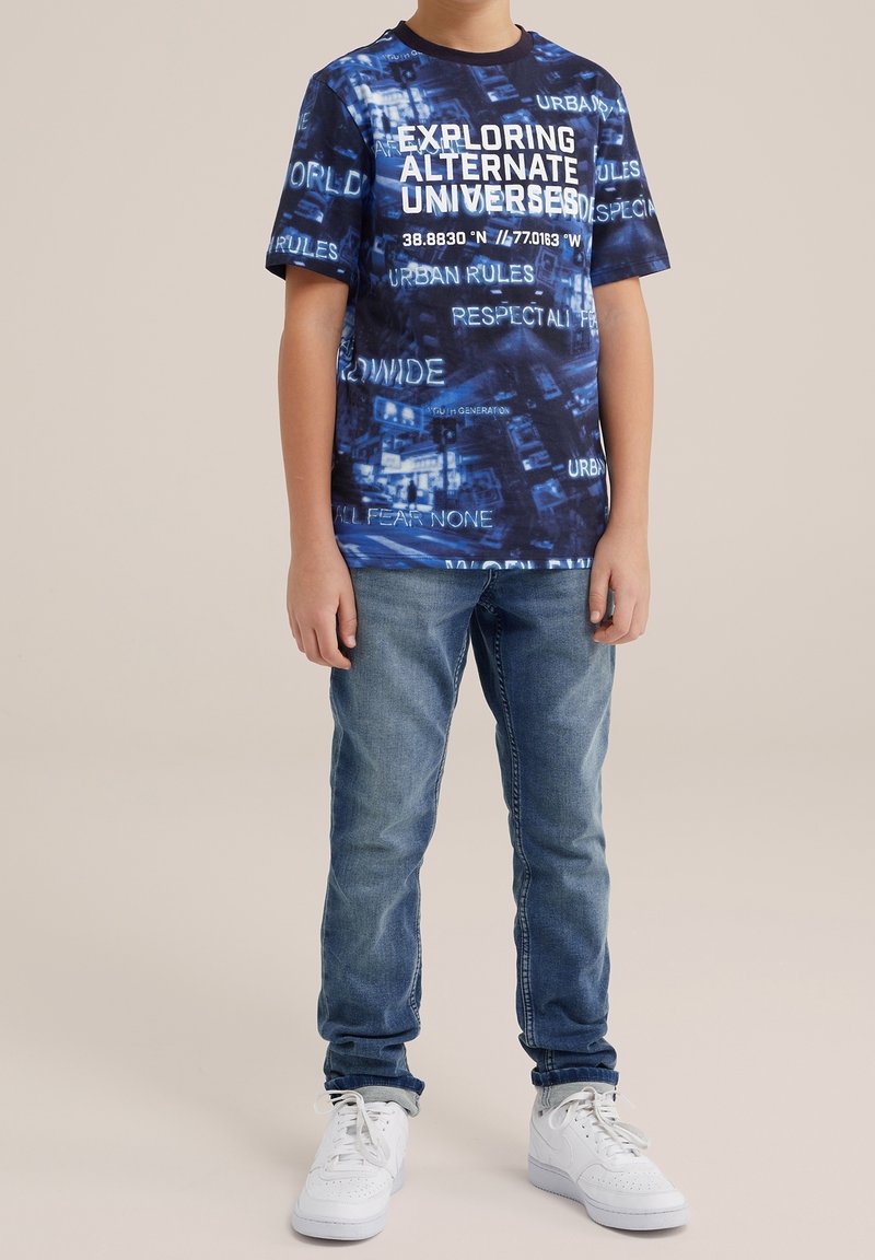 T-shirt grafica blu navy con testo bianco a tema urbano, abbinata a jeans azzurri e sneaker bianche. Design semplice con vestibilità casual.