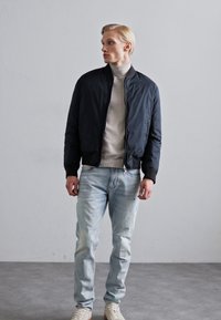 Svart bomberjacka med dragkedja fram, ribbade ärmslut och en jämn textur, bärs över en ljusgrå polotröja och ljusblå jeans.