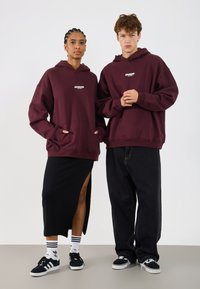 Hanorace bordo cu buzunare frontale, logo pe piept, asociate cu o fustă neagră cu fanta laterală și blugi negri largi, purtate cu adidași negri.