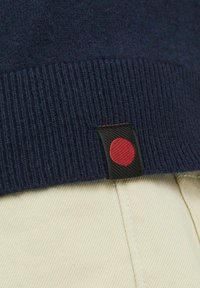 Pulover din lână de culoare bleumarin, cu manșete cu nervuri, având o etichetă neagră cu un accent roșu. Pantaloni de culoare deschisă vizibili dedesubt.