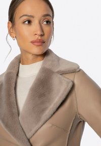 Hellbraune Lederjacke mit einem plüschigen beigefarbenen Fellkragen und einem strukturierten Design. Darunter trägt sie einen gerippten weißen Pullover.