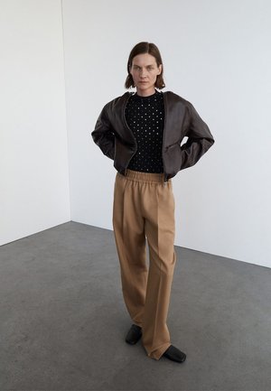 Braune Lederbomberjacke, schwarzer Polka-Dot-Pullover, beigefarbene, plissierte Hose und schwarze Slipper. Minimalistisches Design, glatte Texturen.