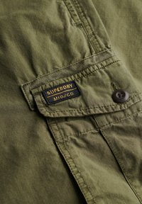 Tessuto di camicia verde oliva con tasca frontale, caratterizzato da un'etichetta "SUPERDRY" nera e gialla, e un dettaglio con bottone marrone. Finitura testurizzata.